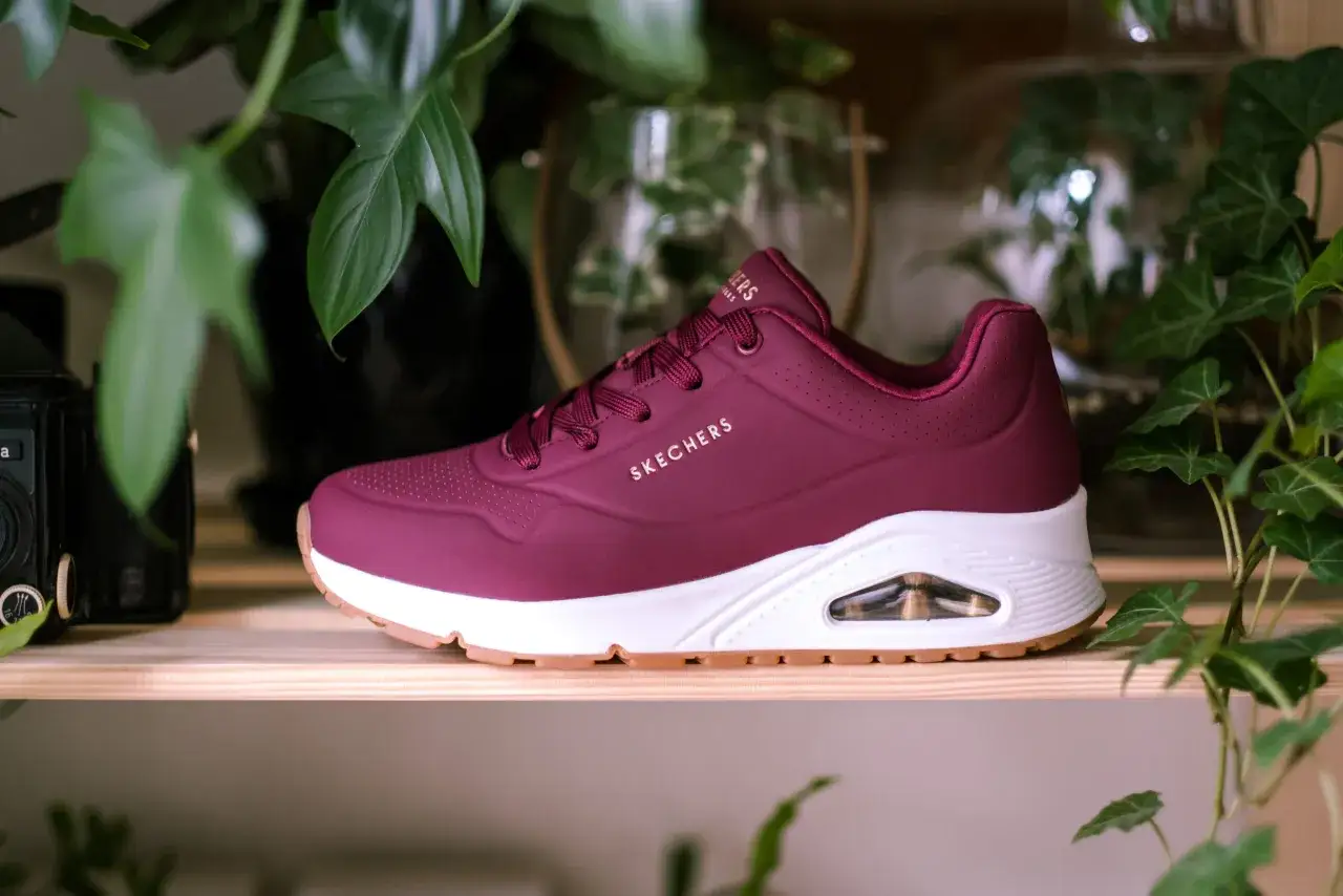 Bordowy but Skechers z białym spodem i brązową podeszwą, otoczony zielonymi liśćmi. Dowiedz się, jak wyczyścić sznurówki, aby buty wyglądały jak nowe.