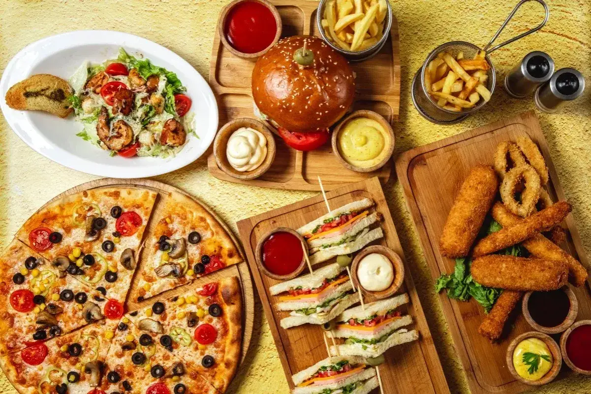 Obfity stół z jedzeniem fast food: pizza, burgery, frytki, kanapki, sałatka i panierowane przekąski.