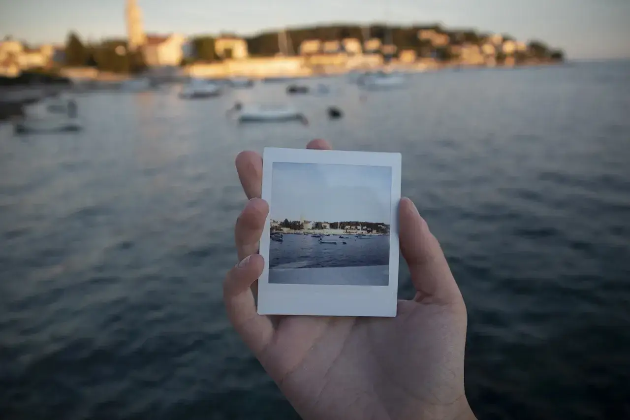 Dłoń trzyma zdjęcie z widokiem na zatokę z łodziami, jak wywoływanie zdjęć jak z instaxa.