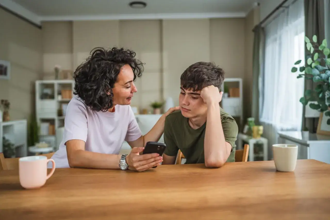 Matka z troską patrzy na telefon syna, zastanawiając się, czy rodzic ma prawo sprawdzać dziecku telefon.