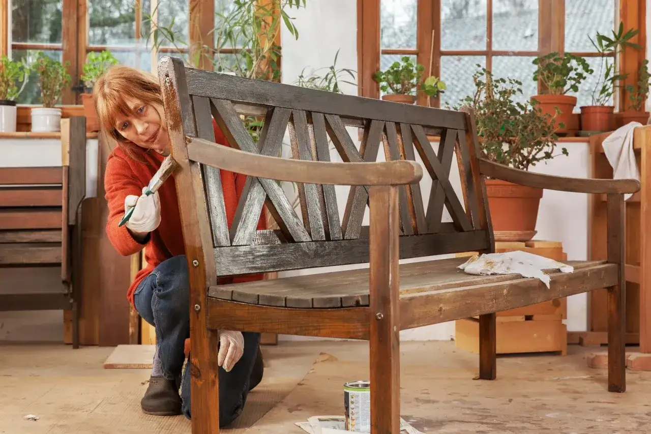 Kobieta maluje drewnianą ławkę ogrodową. Zastanawiasz się, jakie drewno na meble ogrodowe będzie najlepsze?
