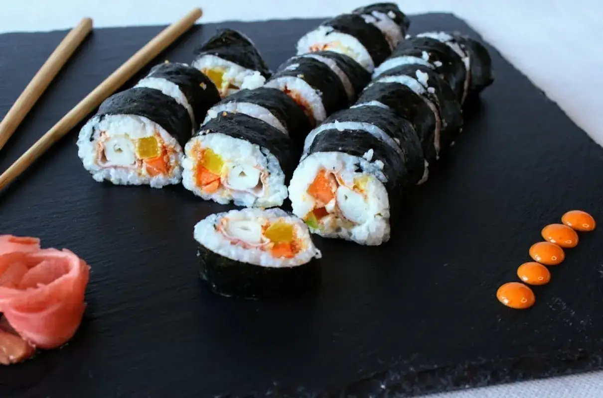 Roladki sushi z surimi, ryżem i warzywami na czarnej desce, obok pałeczki i różowy imbir.