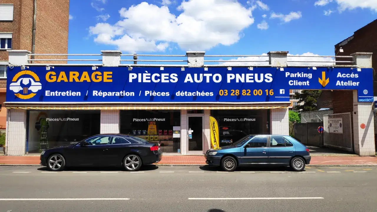 Garage avec enseigne bleue et jaune, deux voitures garées devant, sous un ciel bleu avec des nuages.