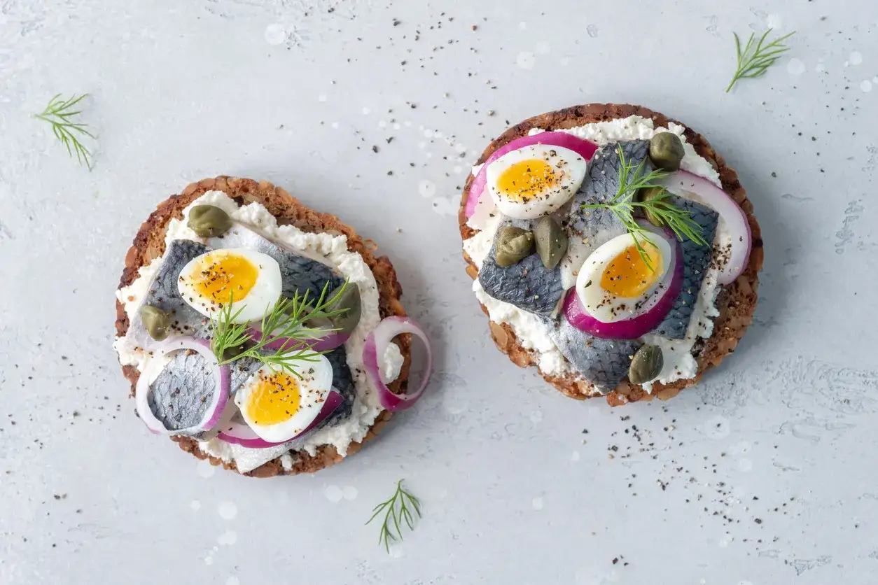 Dwa otwarte kanapki z pastą serową, śledziem, jajkiem przepiórczym, cebulą i kaparami. Idealna przystawka przed obiadem.