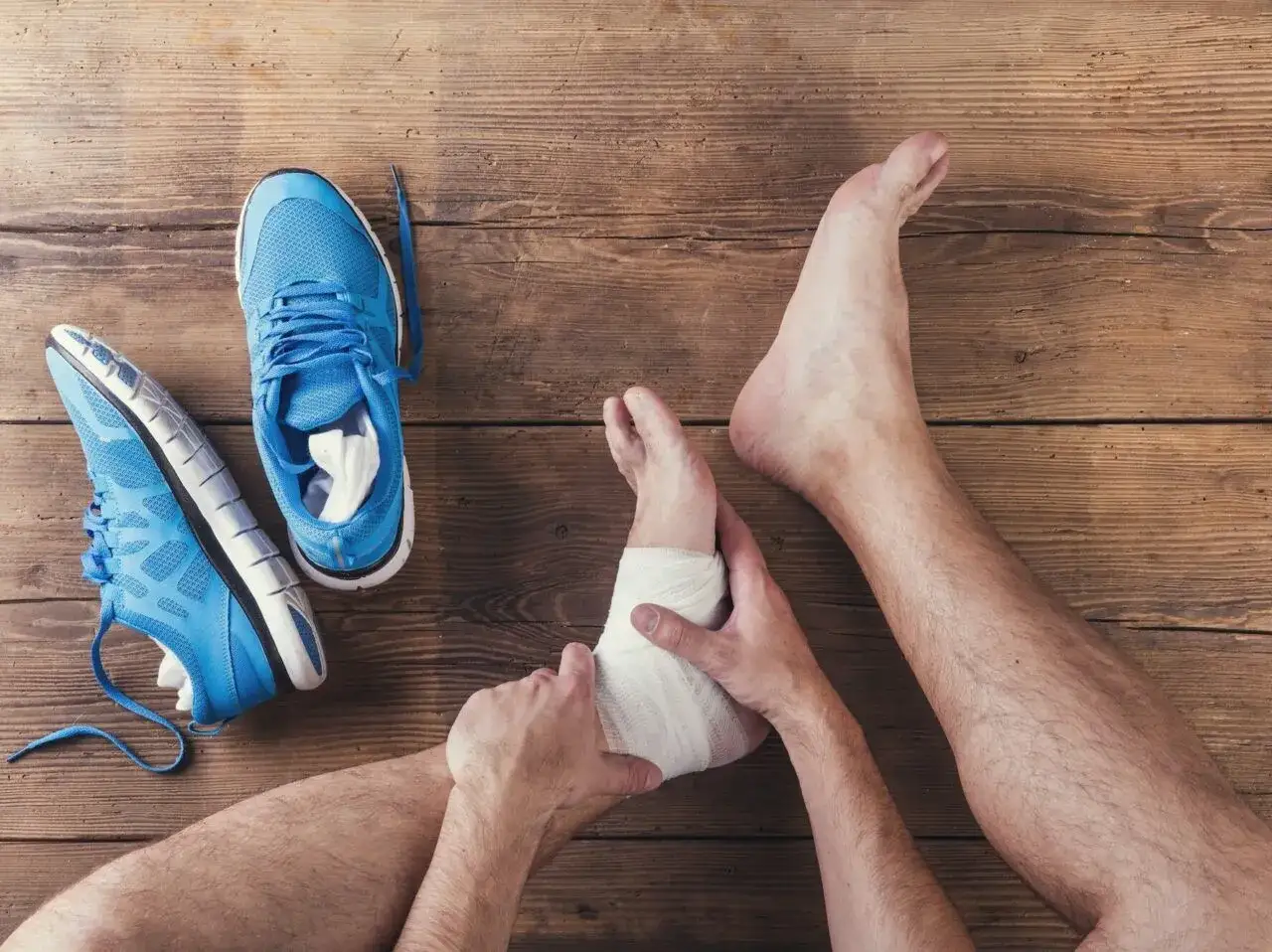 Opatrywanie skręconej kostki bandażem, obok leżą niebieskie buty sportowe. Dowiedz się, jak pozbyć się opuchlizny po skręceniu kostki.