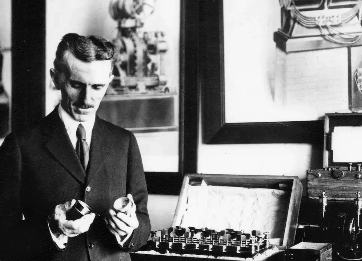 Nikola Tesla w garniturze trzymający dwa metalowe elementy, obok niego aparatura.