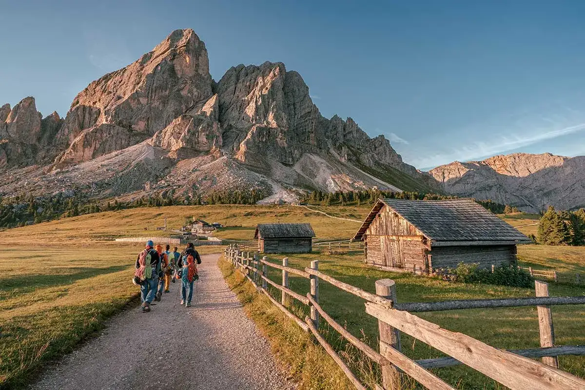 Piękna fotografia krajobrazowa Dolomitów. Grupa turystów wędruje ścieżką obok drewnianych chat, pod majestatycznymi szczytami górskimi.