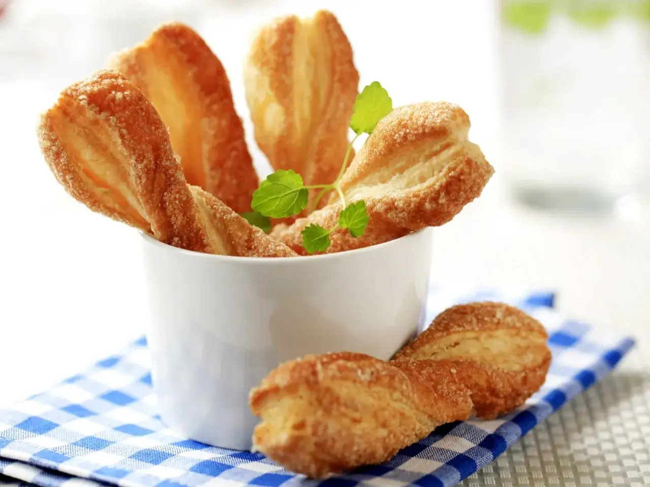 Chrupiące paluchy z ciasta francuskiego posypane cukrem, podane w białym naczyniu na niebiesko-białej kratce.