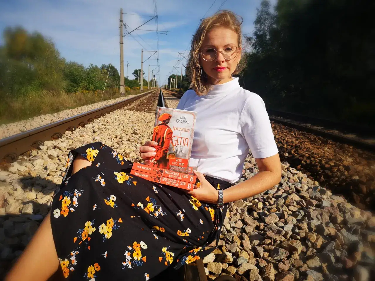 Kobieta z książką "Nauczycielka z Getta" siedzi na torach. Recenzja książki gwiazd naszych win po angielsku.