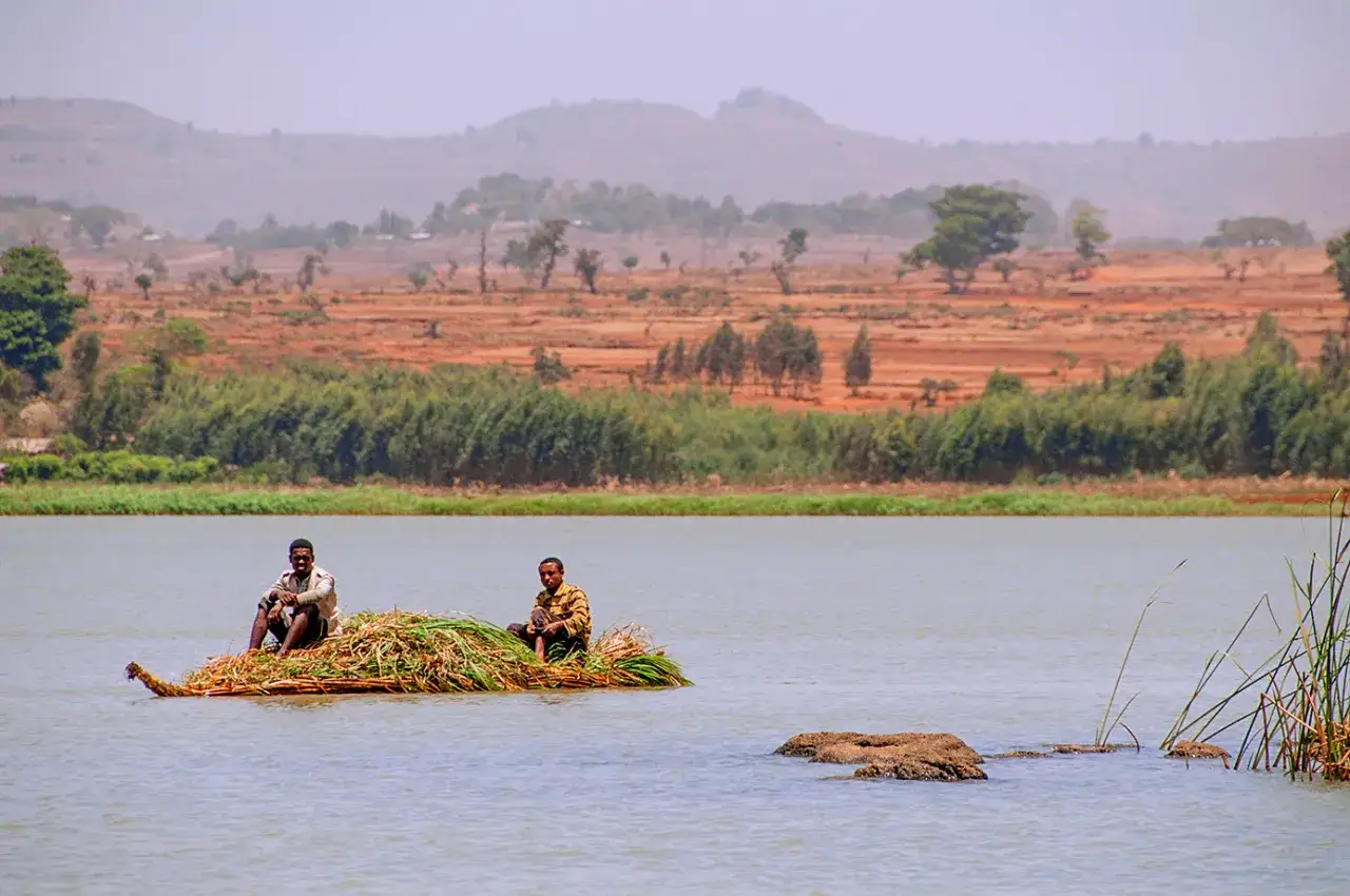 Dwóch mężczyzn na tratwie z trawy na spokojnym jeziorze w Etiopii. W tle suche, pofałdowane wzgórza i zieleń.