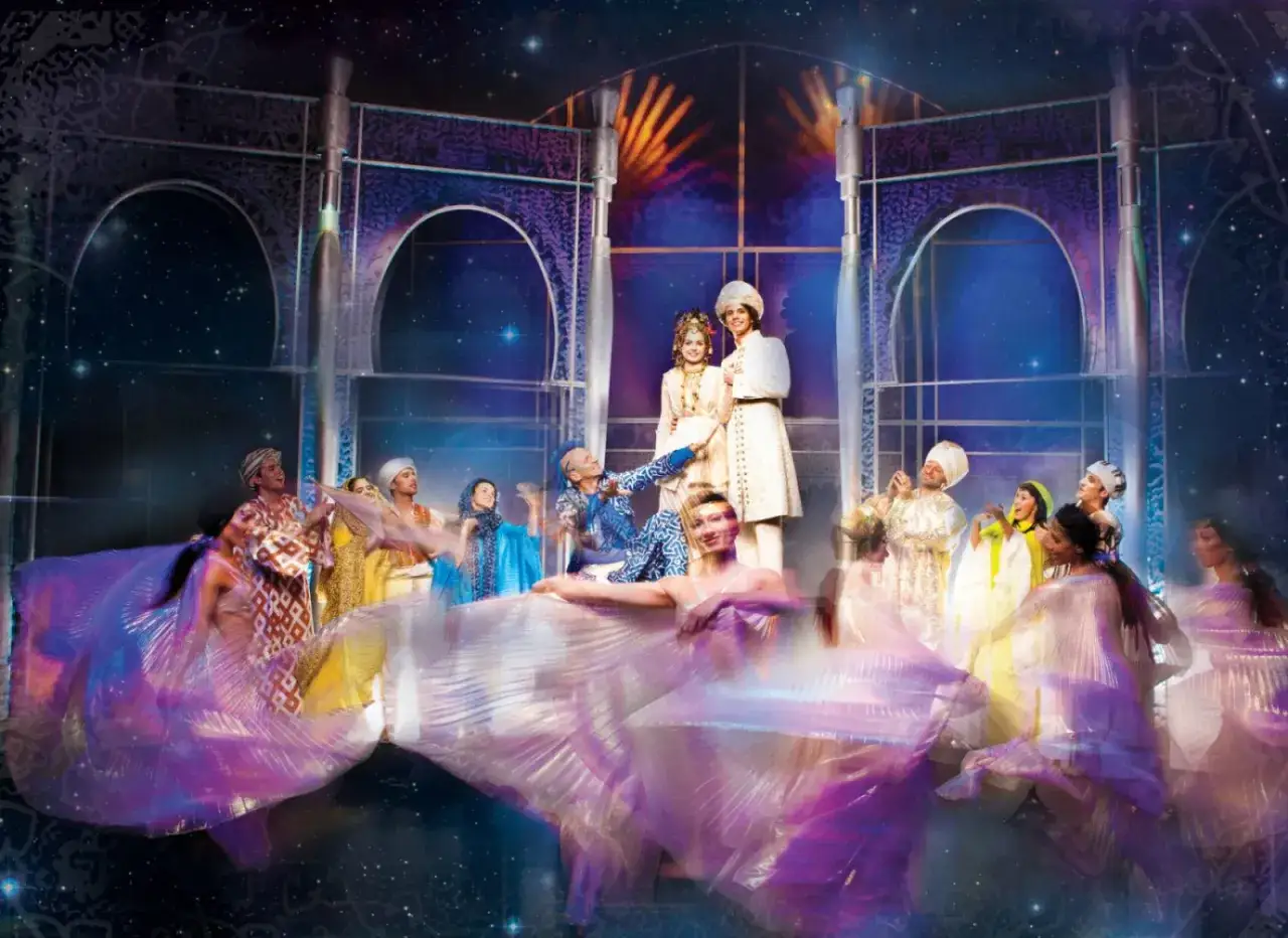 Scena z musicalu "Aladyn" w teatrze w Warszawie. Tancerze w barwnych strojach i księżniczka z księciem na scenie.
