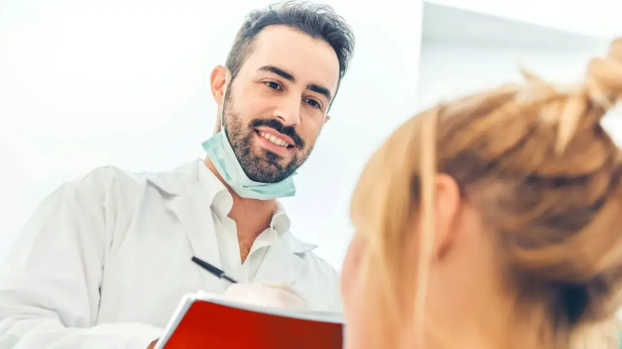 Uśmiechnięty dentysta w białym fartuchu i masce na szyi rozmawia z pacjentką, trzymając notatnik.
