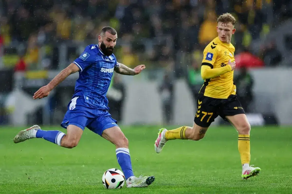 Piłkarz w niebieskiej koszulce kopie piłkę, a za nim biegnie zawodnik w żółtej.