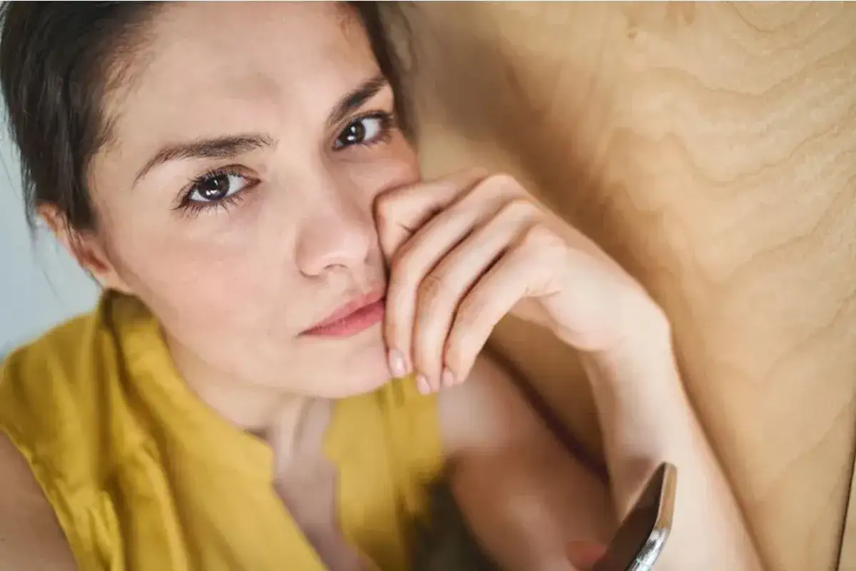 Zbliżenie na twarz kobiety w żółtej bluzce, opierającej głowę na dłoni, z telefonem w drugiej ręce.