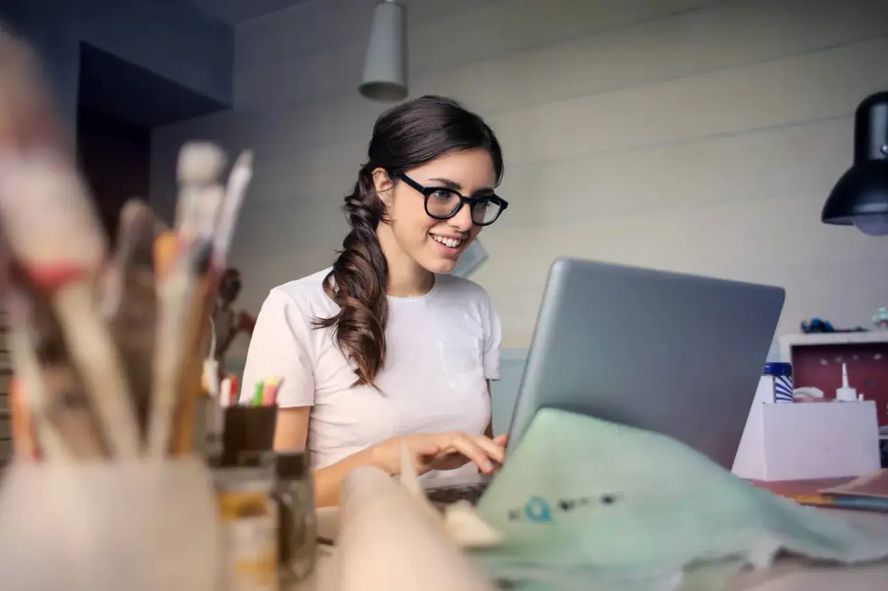 Uśmiechnięta kobieta w okularach pracuje przy laptopie, realizując swoją pracę dodatkową z domu.
