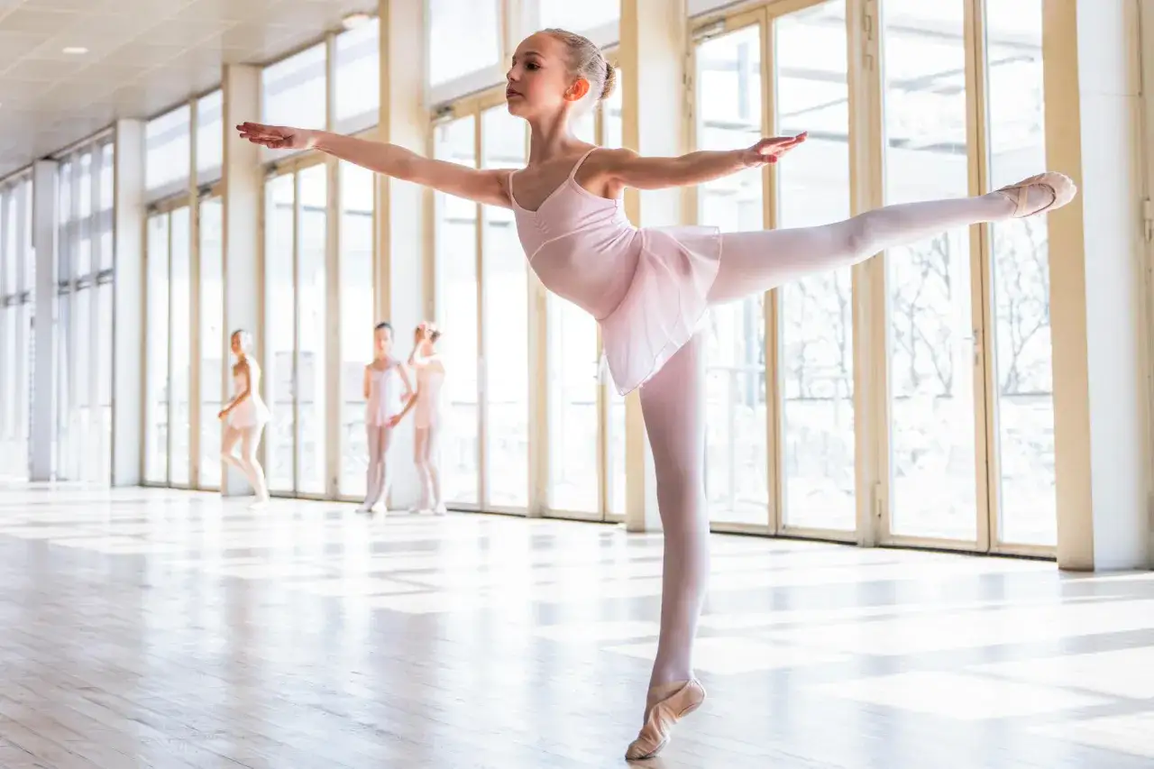 Młoda baletnica w różowej sukience wykonuje eleganckie pozycje baletowe. W tle inne tancerki.