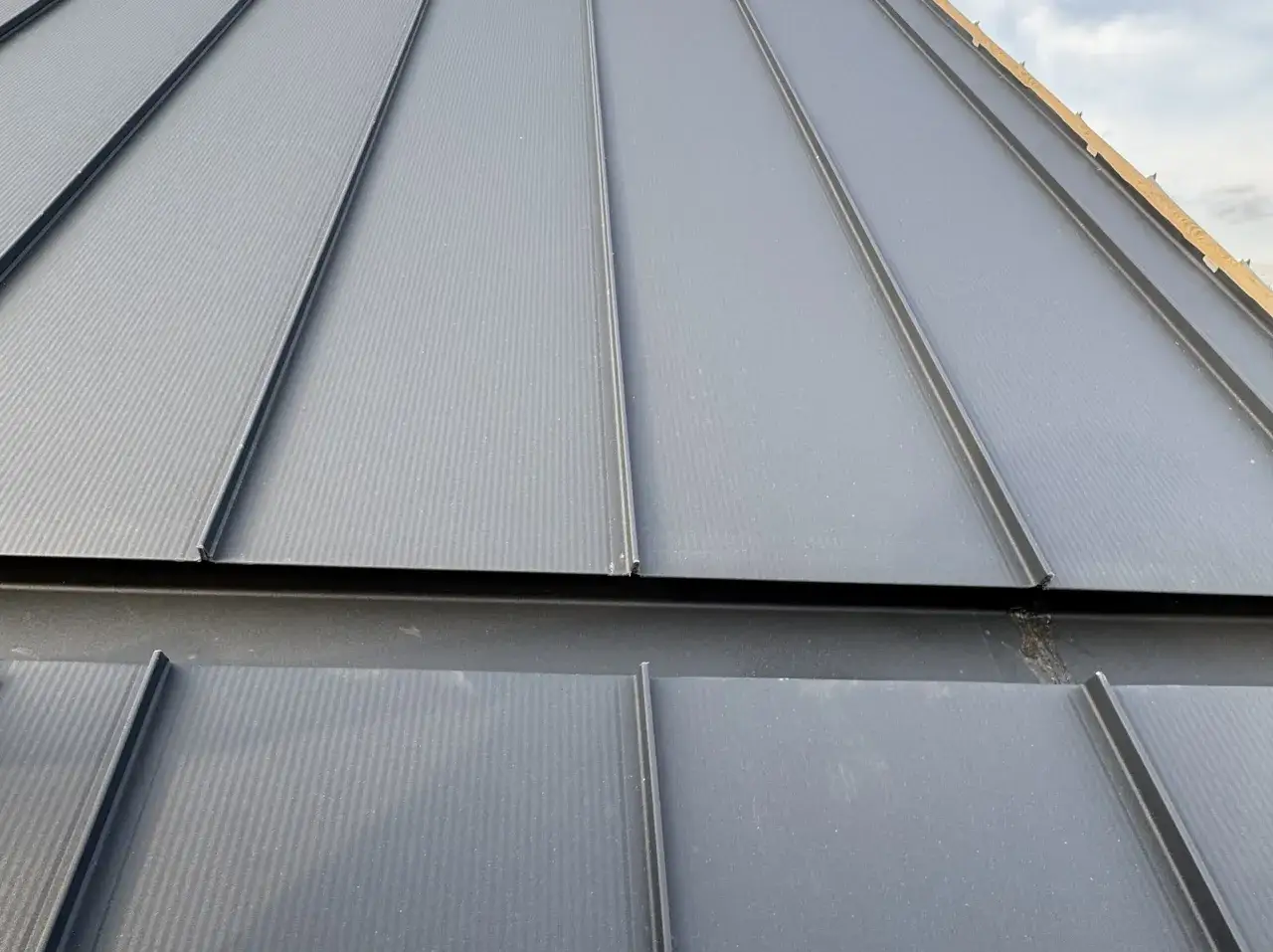 Szara blacha na rąbek stojący z widocznymi wadami montażu, np. nieszczelnościami na połączeniach.