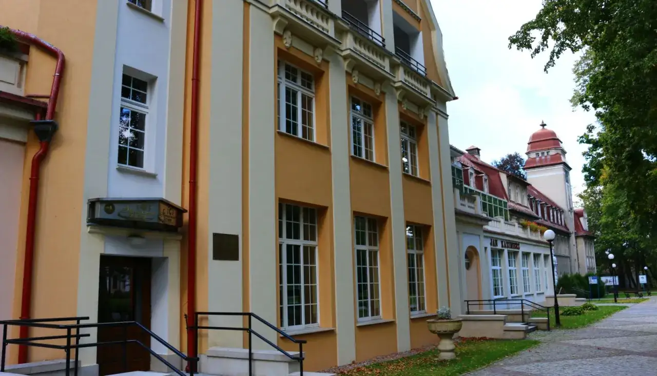 Sanatorium Jan Kazimierz w Dusznikach-Zdroju, z charakterystyczną żółto-kremową fasadą i czerwonym dachem, otoczone zielenią.