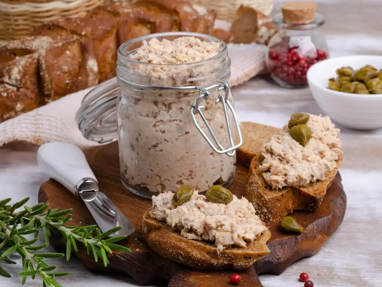 Słoik pełen pysznej pasty z tuńczyka, gotowej do rozsmarowania na chlebie. Na desce kromki z pastą i kaparami, obok gałązka rozmarynu.