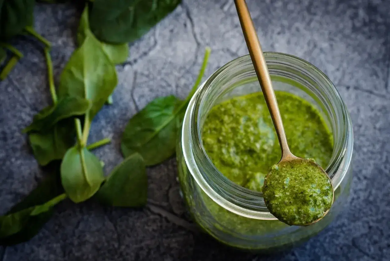 Słoik z zielonym pesto i łyżka z porcją sosu, obok liście szpinaku.