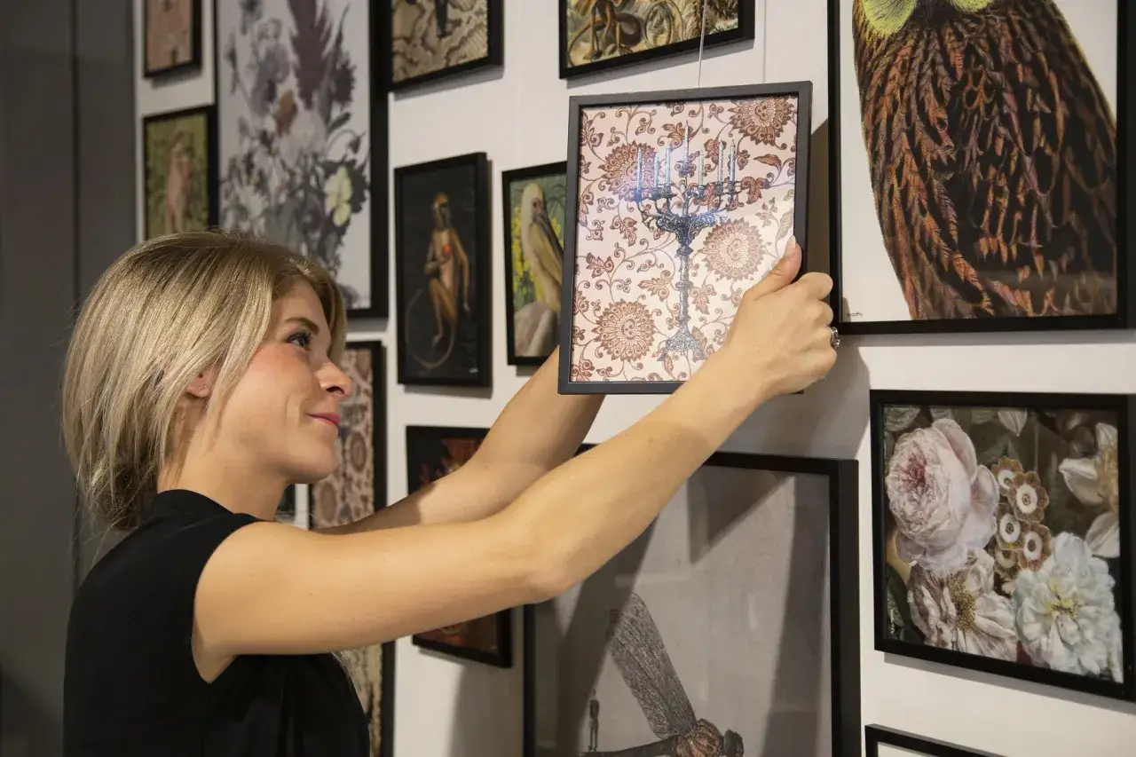 Kobieta wiesza obraz na ścianie. W tle inne oprawione grafiki.