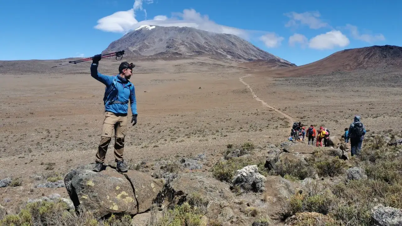 Wspinacz na Kilimandżaro z kijkami, w tle szlak i grupa turystów. Temperatura na szczycie może być mroźna.