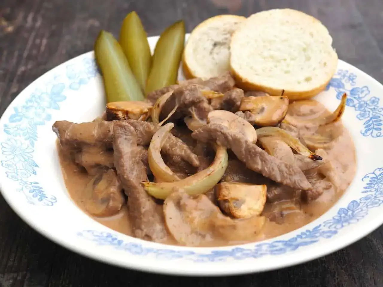 Tradycyjny strogonow z wołowiny z pieczarkami i cebulą, podany z ogórkami i bułeczkami. Pyszny przepis na obiad.