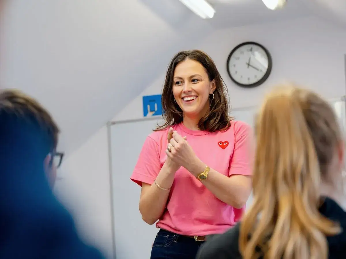 Uśmiechnięta kobieta w różowej koszulce z sercem, stojąca przed uczniami.