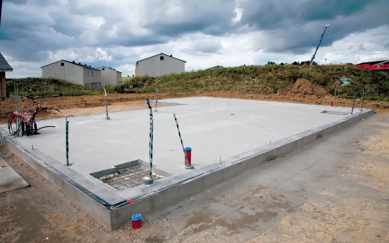 Budowa płyty fundamentowej w trakcie. Betonowa płyta z wystającymi rurami i kablami, w tle domy w budowie.