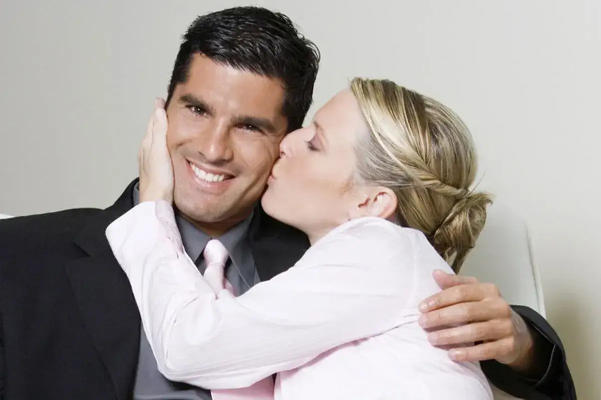 Kobieta całuje w policzek uśmiechniętego mężczyznę w garniturze. To mogą być zakochany szef objawy, gdy pracownica okazuje mu sympatię.