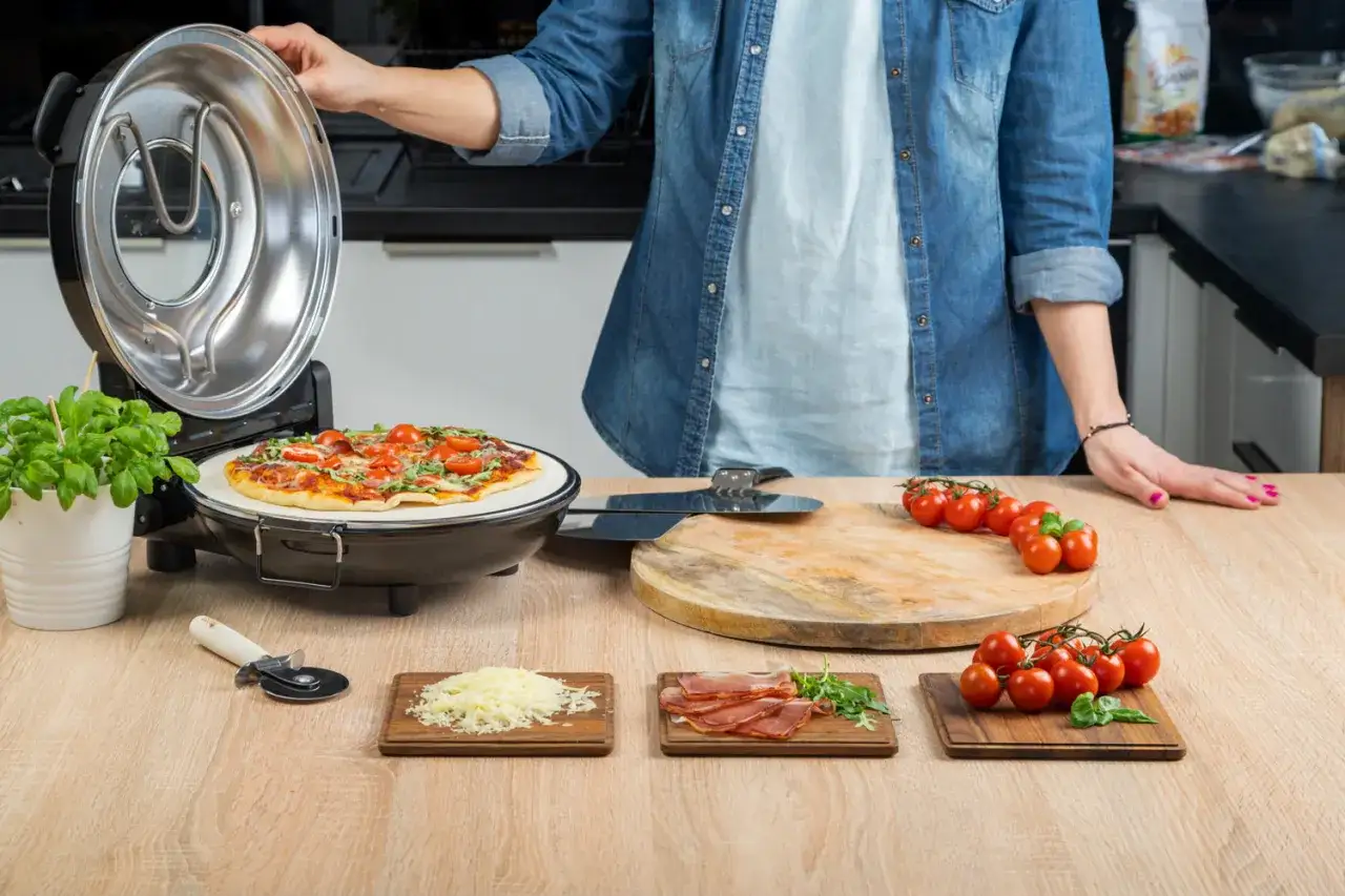 Otwarta opiekacz do pizzy, na kamieniu piec pizza z pomidorkami i bazylią. Obok deska z pomidorkami, startym serem i szynką.