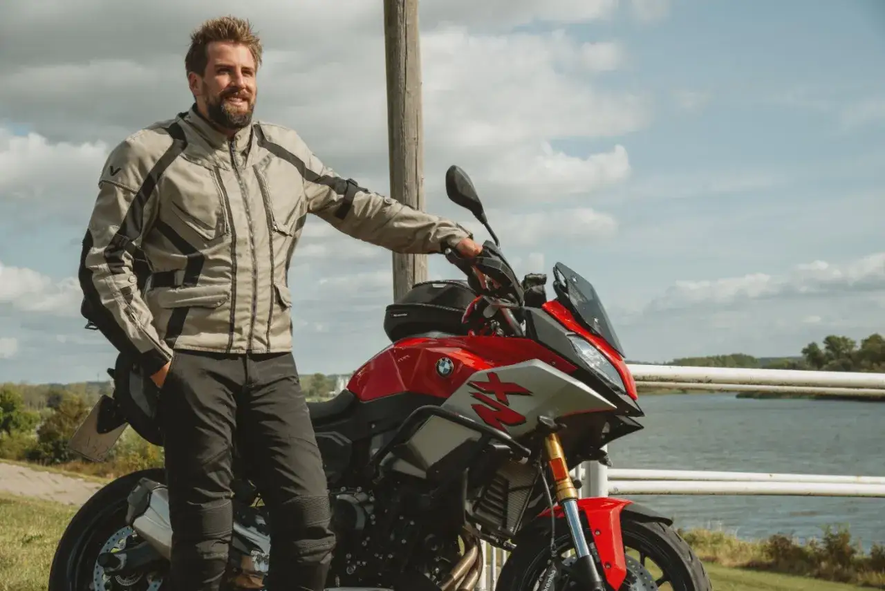 Uśmiechnięty motocyklista w kurtce od polskiego producenta odzieży motocyklowej, oparty o czerwoną maszynę nad jeziorem.