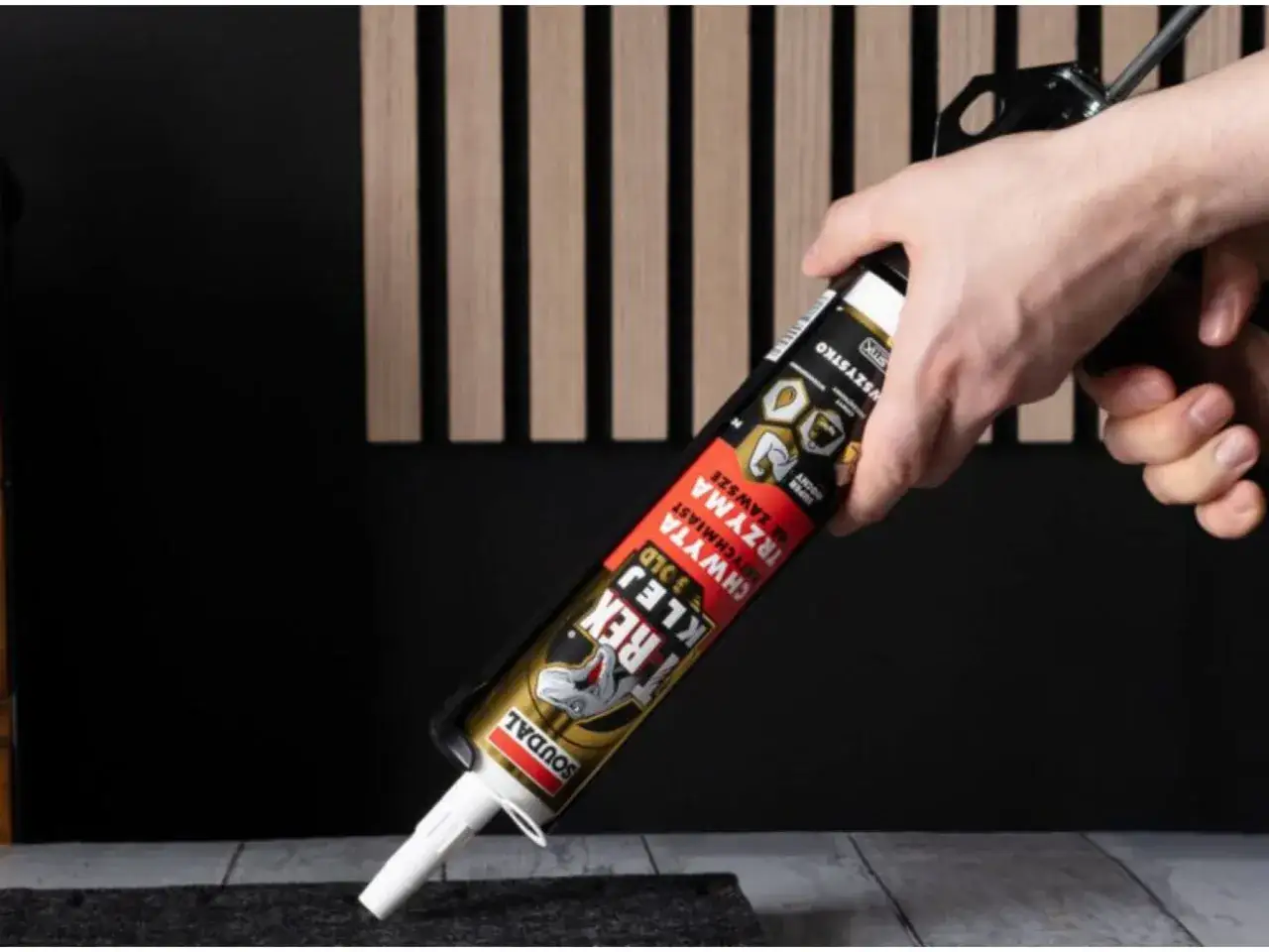 Dłoń trzymająca pistolet z tubą kleju montażowego na tle lameli.