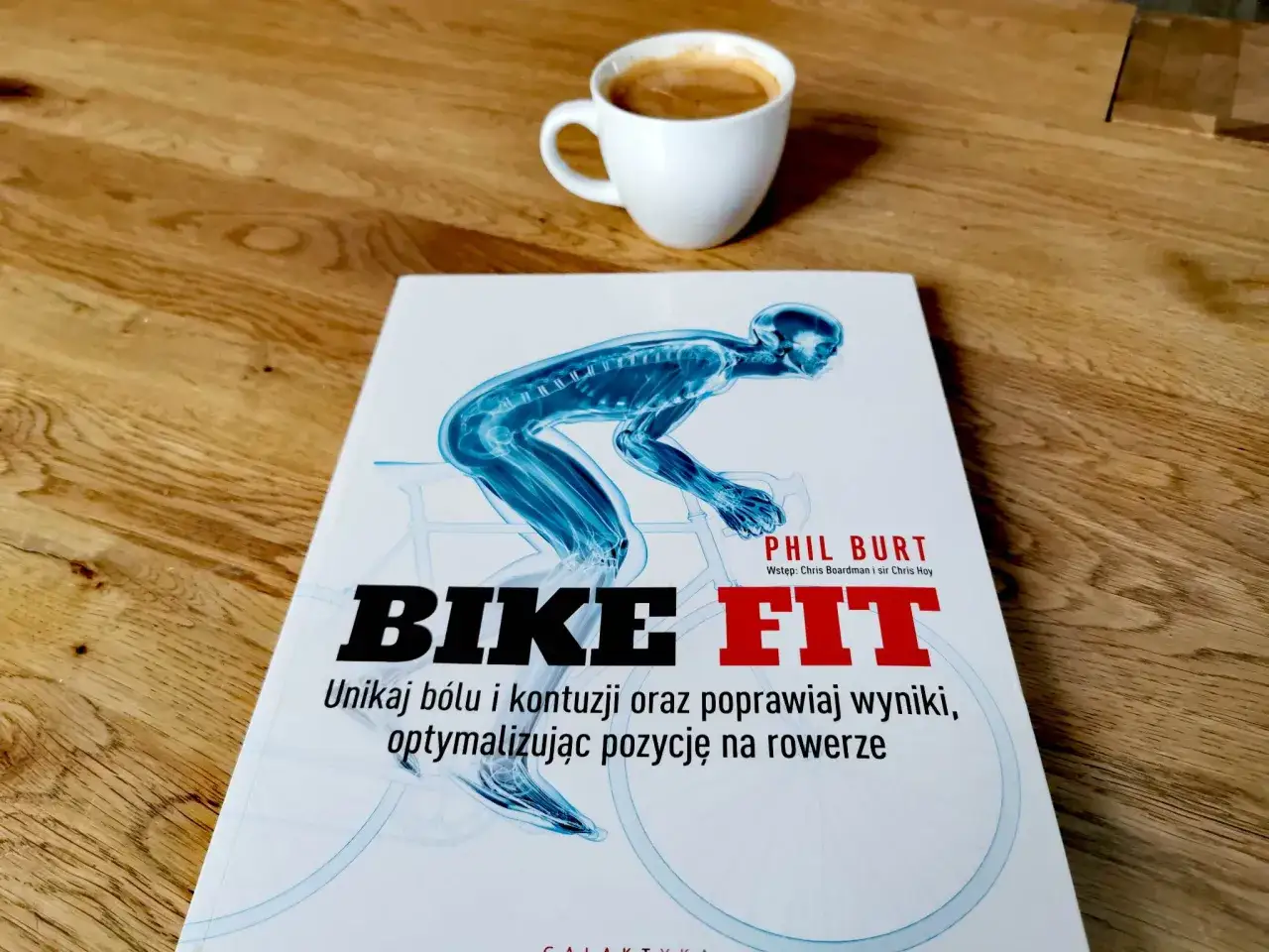 Książka "Bike Fit" z anatomicznym schematem rowerzysty i filiżanką kawy na drewnianym blacie.
