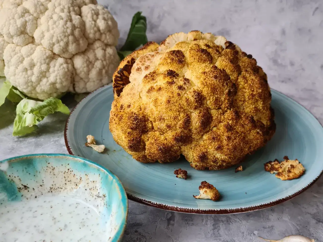 Złocisty, pieczony kalafior na talerzu, obok świeży i miseczka sosu. Idealny pomysł co do kalafiora na obiad.