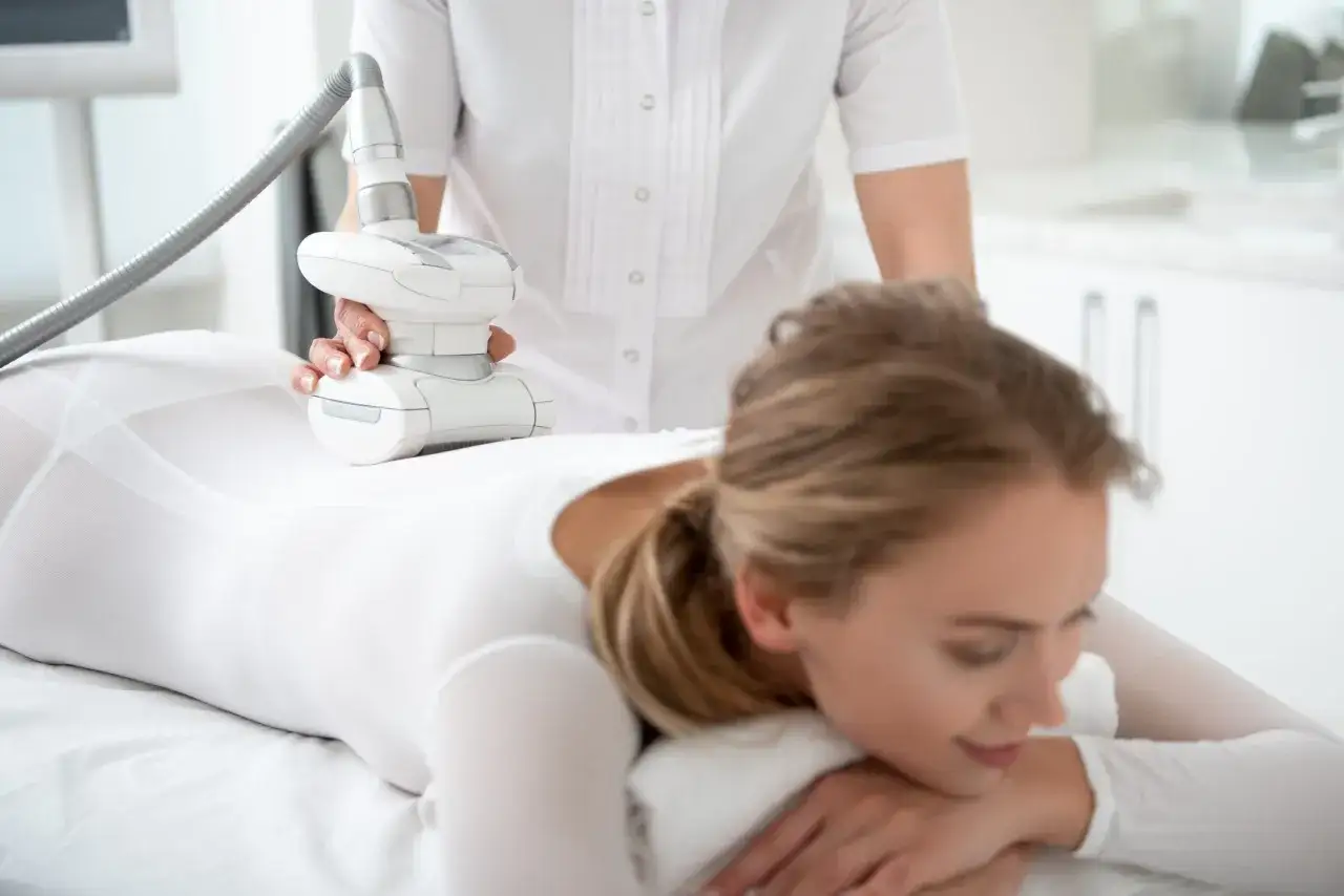 Kobieta w białym stroju poddaje się zabiegowi endermologii LPG. Widoczne są ręce terapeuty trzymające głowicę urządzenia, które masuje nogę pacjentki. Efekty zabiegu są obiecujące.