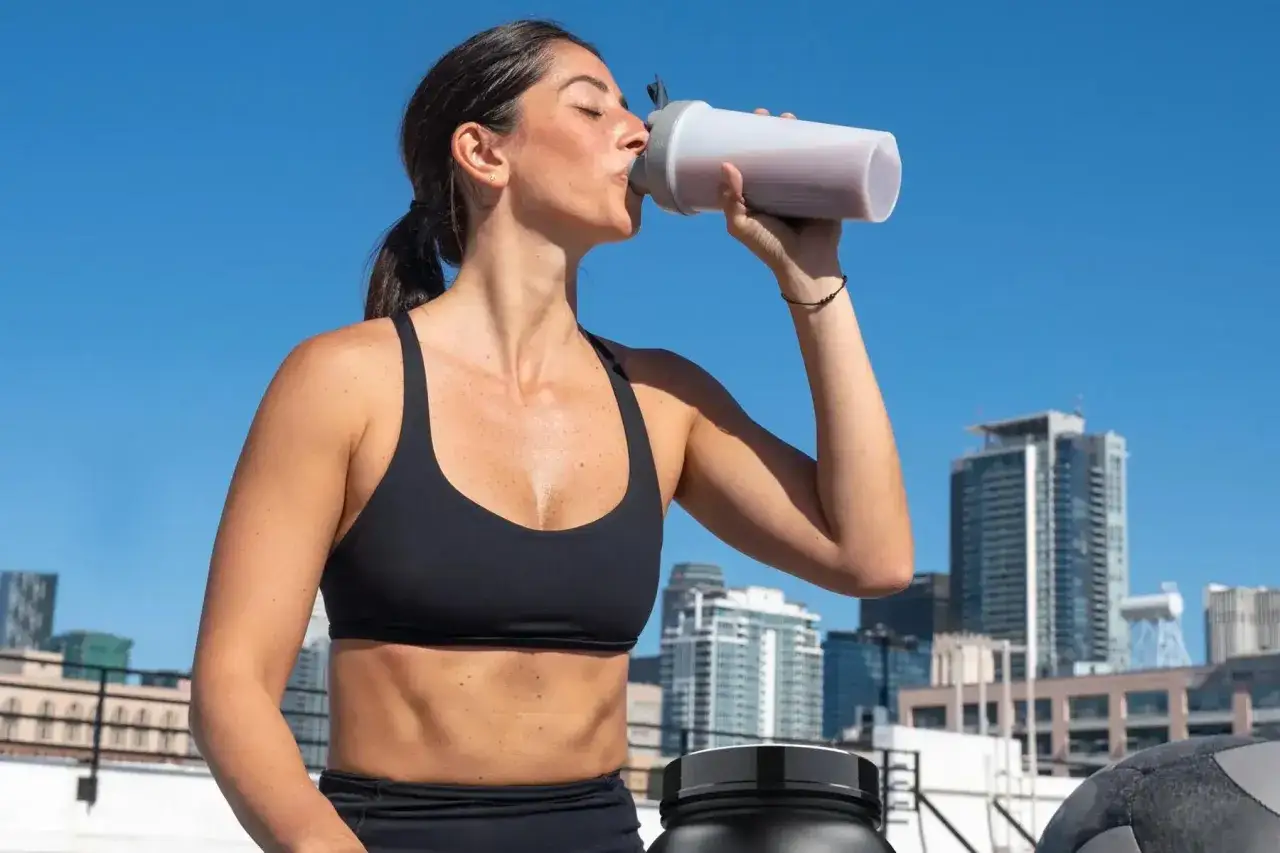 Kobieta w sportowym topie pije odżywkę białkową z bidonu na tle miasta.