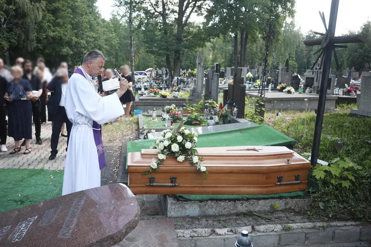 Wojciech Magnuski żegna zmarłego na cmentarzu. Ksiądz w białej szacie czyta z księgi przy trumnie ozdobionej białymi różami.