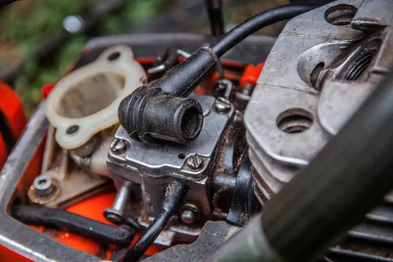 Zbliżenie na silnik kosiarki spalinowej: świeca zapłonowa, gaźnik i cylinder.
