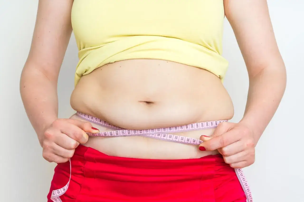 Kobieta mierzy obwód talii centymetrem. Widoczny jest odstający brzuch poniżej pępka, co może świadczyć o nadwadze.
