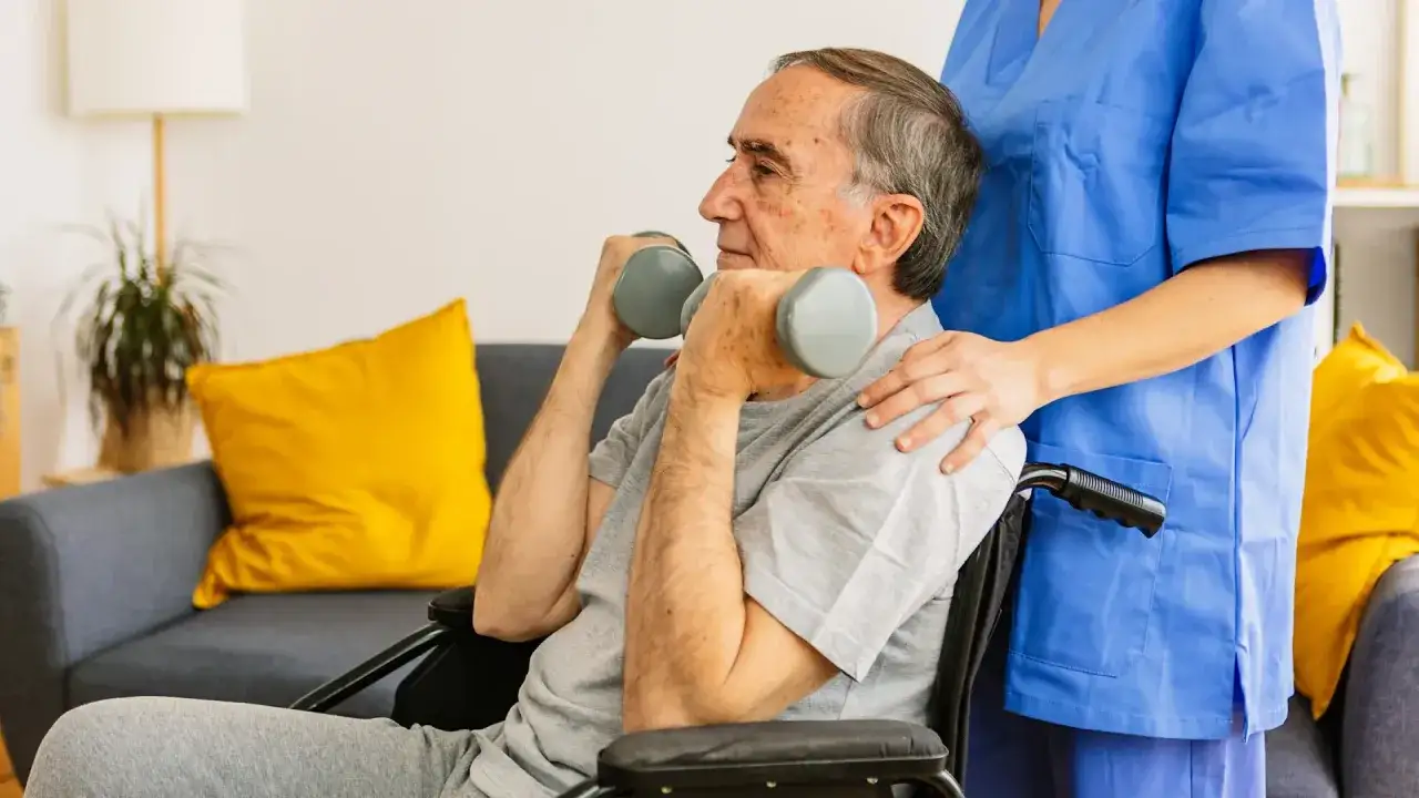 Starszy mężczyzna w wózku inwalidzkim ćwiczy z hantlami, wspierany przez pielęgniarkę.