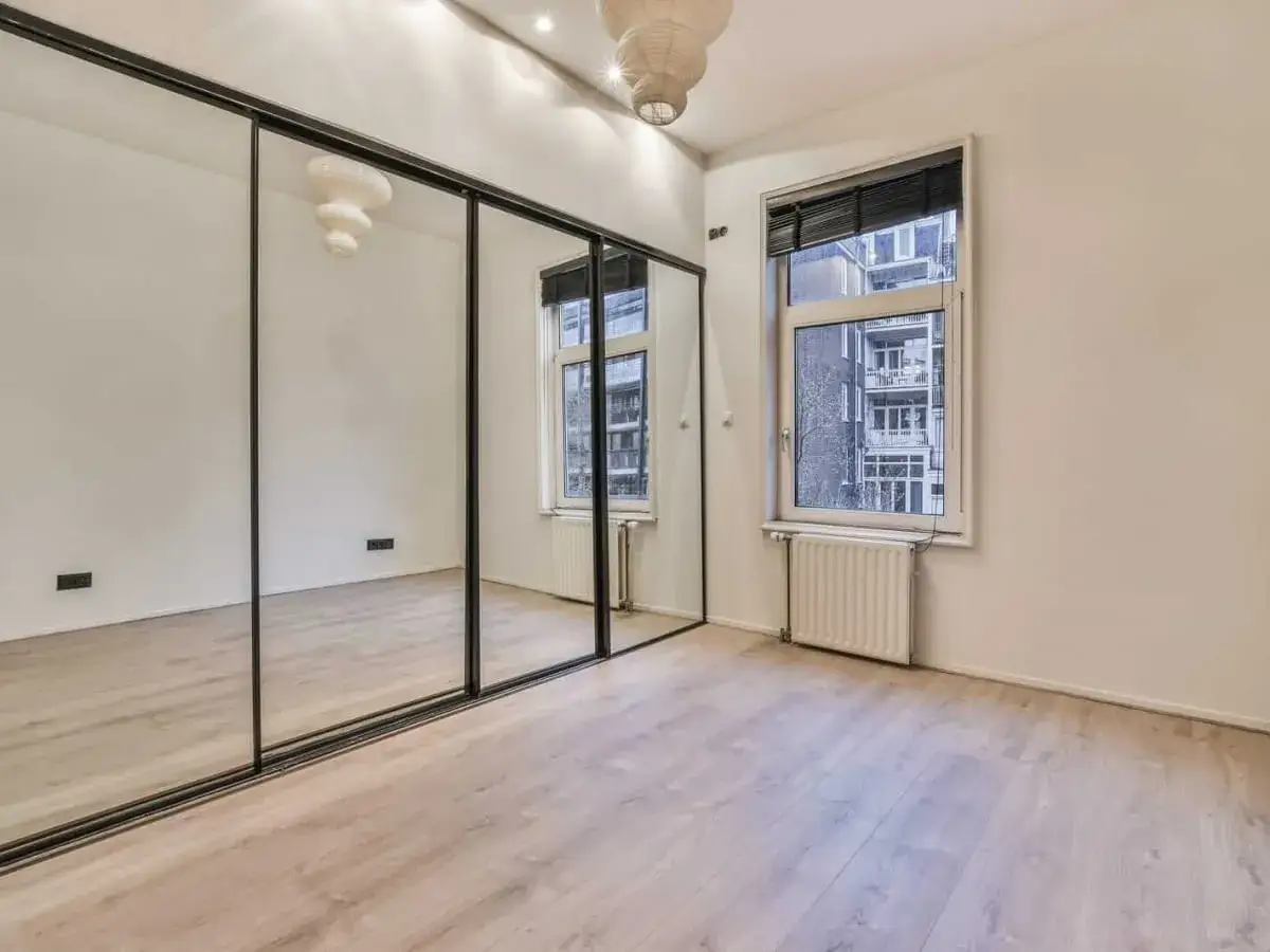 Przestronne wnętrze z dużą, lustrzaną szafą, która optycznie powiększa pokój. Obok okno z widokiem na budynki.