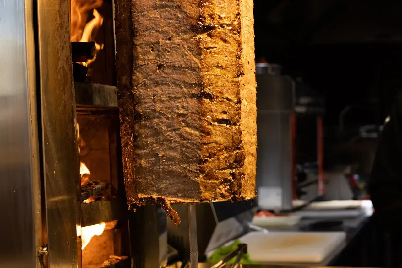Duży pionowy obrotowy grill z mięsem, obok płomienie ognia.