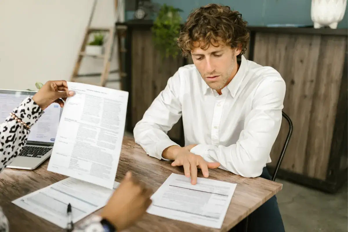 Mężczyzna w białej koszuli analizuje dokumenty z kobietą przy drewnianym stole.