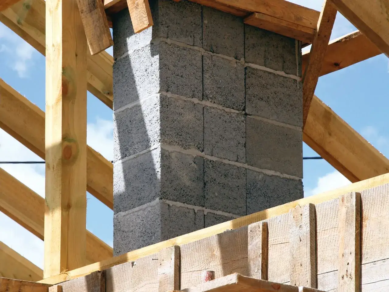 Solidny fundament pod komin systemowy, wznoszony z bloczków betonowych, otoczony drewnianą konstrukcją dachu.