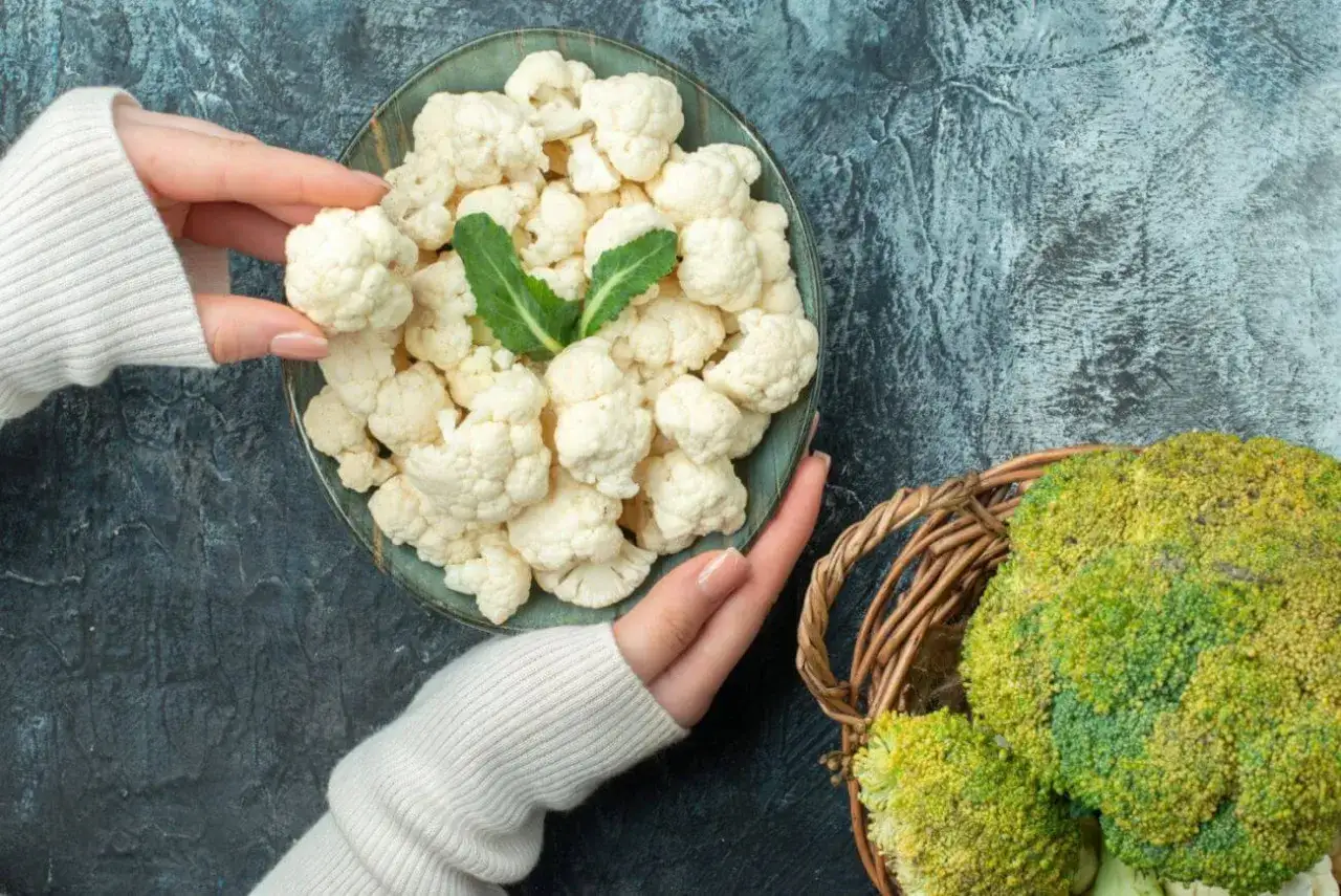 Ręce trzymające różyczki kalafiora nad talerzem, obok koszyk z brokułami. Zastanawiasz się, czy można mrozić kalafior? Tak, można!