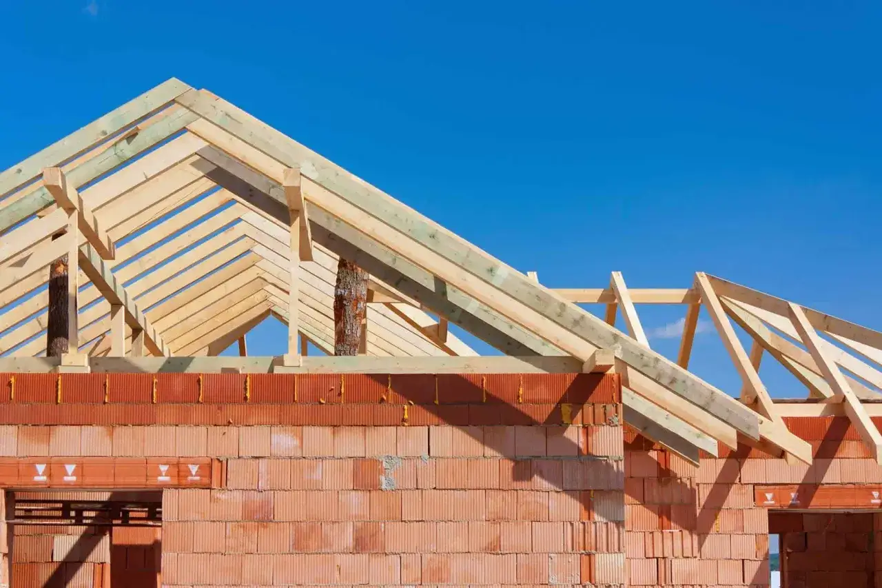 Budowa dachu dwuspadowego w toku. Drewniany szkielet więźby dachowej wznosi się nad ścianami z czerwonej cegły.