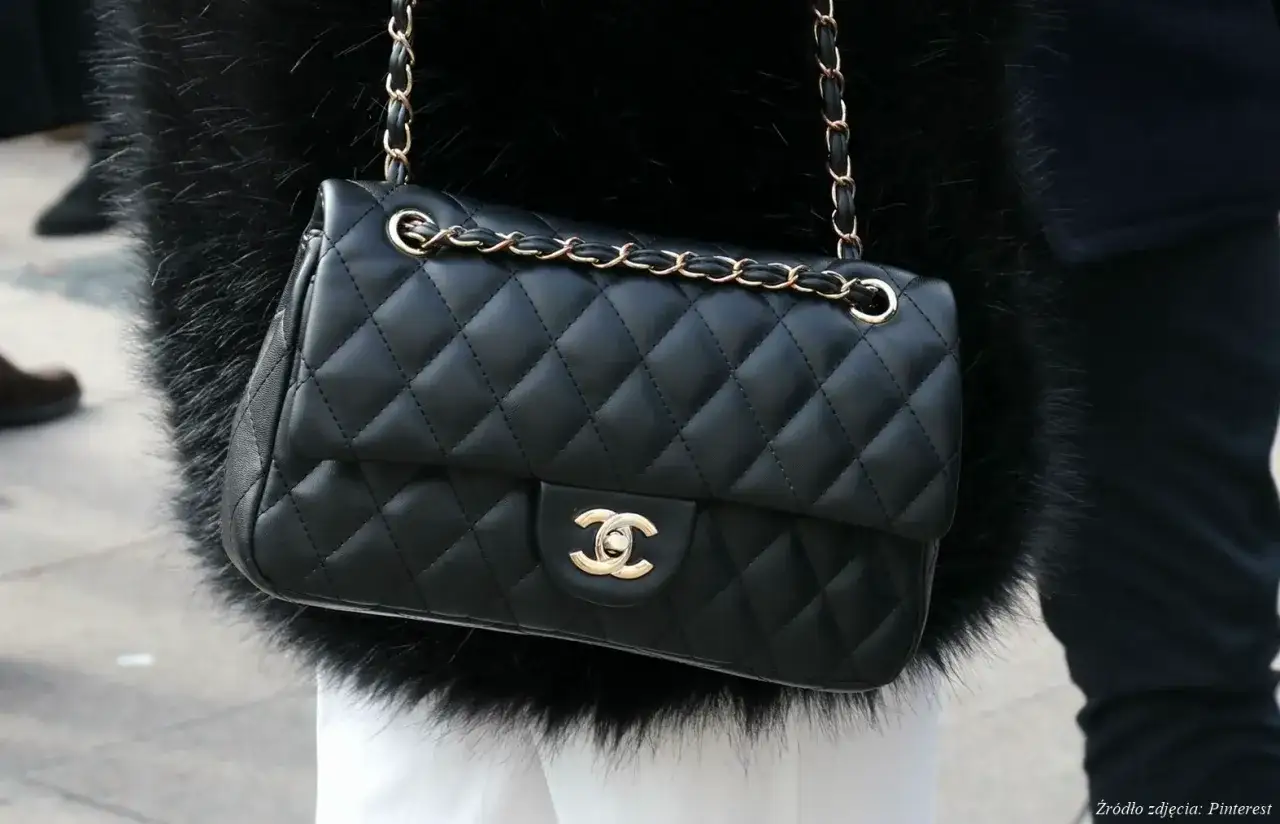 Elegancka, pikowana torebka chanel czarna z złotym łańcuszkiem, noszona na czarnym futrze.
