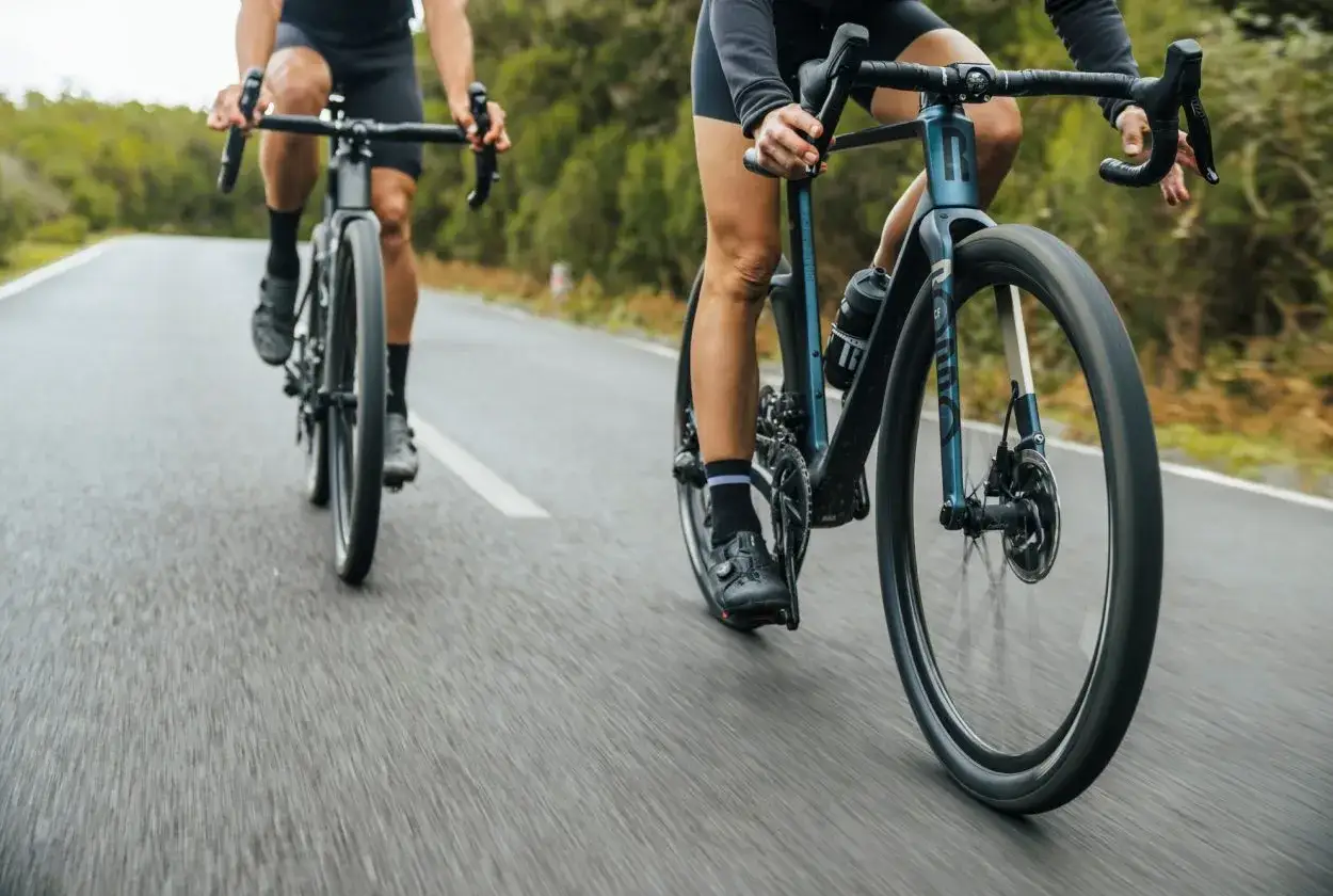 Dwie osoby na rowerach szosowych ROMDO RATT pędzą po asfaltowej drodze wśród zieleni.