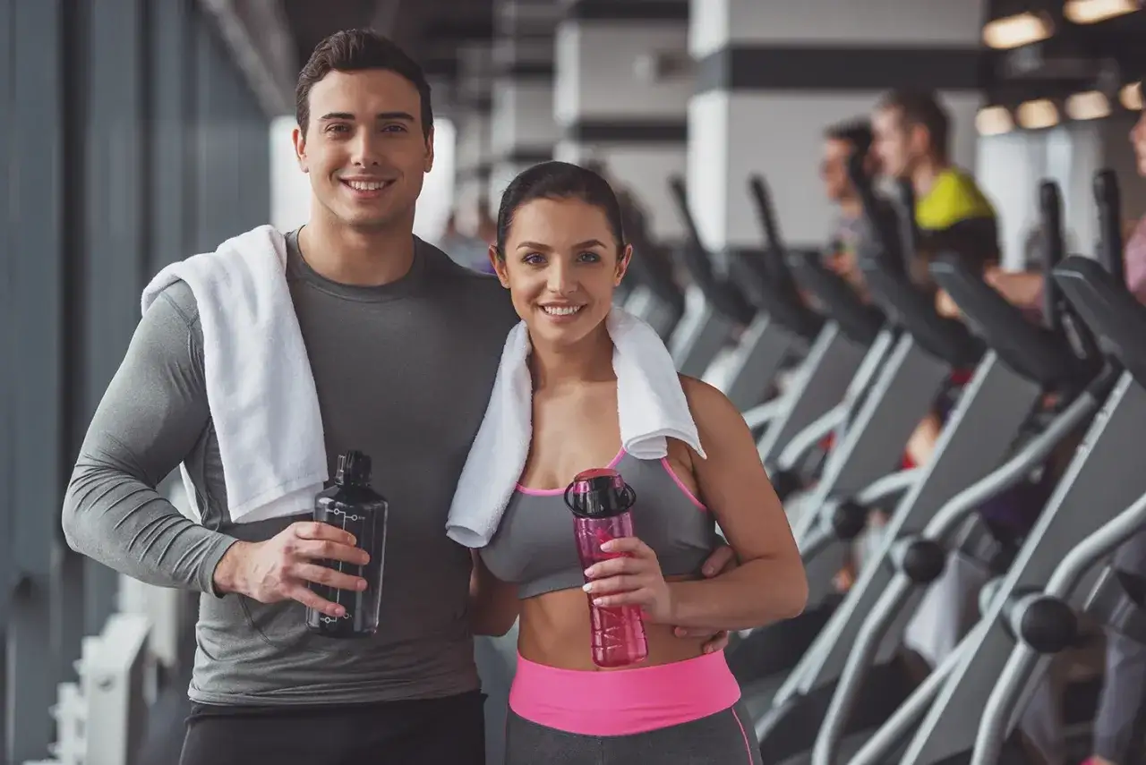 Szczęśliwa para w siłowni, gotowa na trening. Ich uśmiechy i butelki z wodą świadczą o determinacji do poprawy wydolności.